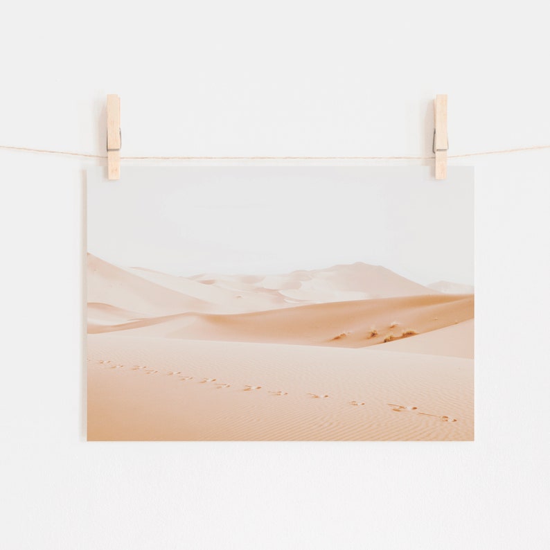 May include: A photograph of a desert landscape with sand dunes and footprints in the sand. The sky is a pale blue and the sand is a warm beige color.
