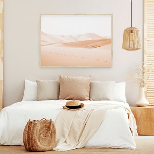 May include: A white bed with a white blanket and a straw hat on top. There are two pillows on the bed, one brown and one white. A brown wicker bag is on the floor next to the bed. A framed picture of a desert landscape hangs on the wall behind the bed.