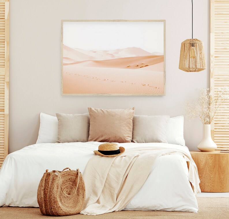 May include: A white bed with a white blanket and a straw hat on top. There are two pillows on the bed, one brown and one white. A brown wicker bag is on the floor next to the bed. A framed picture of a desert landscape hangs on the wall behind the bed.