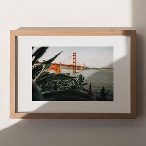 May include: A framed print of the Golden Gate Bridge in San Francisco. The famous orange bridge is set against a body of water, with green foliage in the foreground. The print is presented in a light brown wooden frame, creating a natural aesthetic.