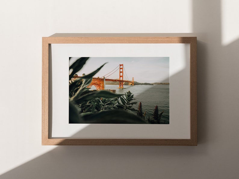 May include: A framed print of the Golden Gate Bridge in San Francisco. The famous orange bridge is set against a body of water, with green foliage in the foreground. The print is presented in a light brown wooden frame, creating a natural aesthetic.