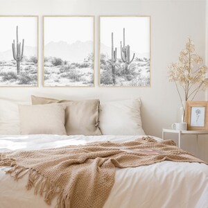 Puede incluir: Tres impresiones en blanco y negro enmarcadas de paisajes des&eacute;rticos con altos cactus. Las impresiones est&aacute;n colgadas en una pared blanca sobre una cama con ropa de cama blanca y una manta de lana marr&oacute;n.