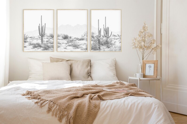 Puede incluir: Tres impresiones en blanco y negro enmarcadas de paisajes des&eacute;rticos con altos cactus. Las impresiones est&aacute;n colgadas en una pared blanca sobre una cama con ropa de cama blanca y una manta de lana marr&oacute;n.
