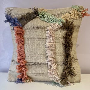May include: Square cushion with a beige woven fabric. The cushion has vertical stripes and is decorated with colourful, textured fringe in shades of blue, peach, green, and brown. A stylish accent for any room.