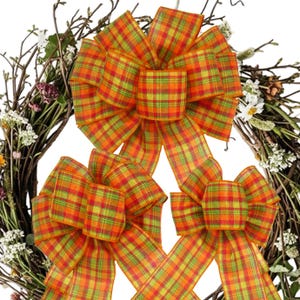 May include: Three decorative bows made of orange, yellow, and green plaid fabric. Each bow features multiple loops and two long, trailing ribbon tails. The bows are arranged in a triangular formation against a background of a rustic twig wreath.