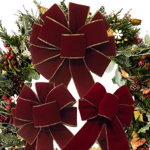 May include: Three deep burgundy velvet bows with metallic gold trim edges. The bows feature multiple loops and long, flowing tails, arranged against a background of a lush green Christmas wreath decorated with pinecones, red berries, and white snowflakes.