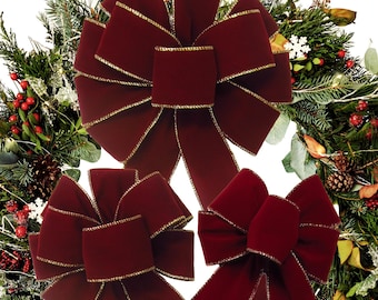 May include: Three deep burgundy velvet bows with metallic gold trim edges. The bows feature multiple loops and long, flowing tails, arranged against a background of a lush green Christmas wreath decorated with pinecones, red berries, and white snowflakes.