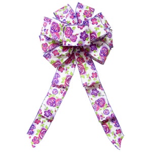 May include: A white bow with purple and pink floral print. The bow is made of satin ribbon and has a wired edge.