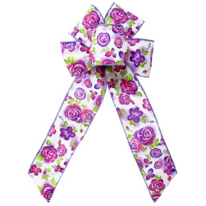 May include: A white bow with a purple and pink floral print. The bow is made of satin ribbon and has a wired edge.