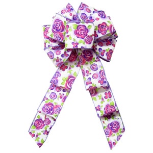 May include: A white bow with purple and pink floral print. The bow is made of satin ribbon and has a wired edge.