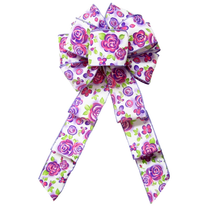 May include: A white bow with purple and pink floral print. The bow is made of satin ribbon and has a wired edge.