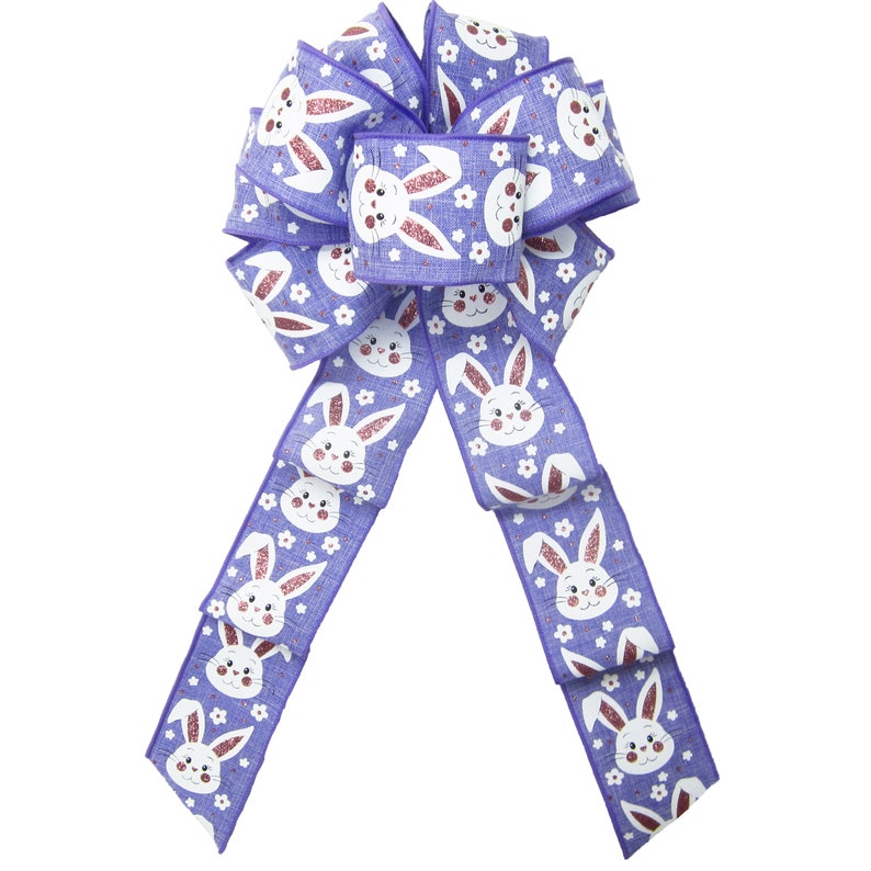 May include: A large Easter bow in periwinkle and white. The bow features a repeating pattern of smiling bunny faces and small white flowers. The bunny ears have a glittery, pink finish. The bow is made of a textured ribbon.