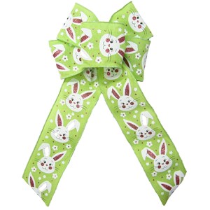 May include: A green bow with white bunny faces and pink flowers. The bow is made of fabric and has a glittery finish.