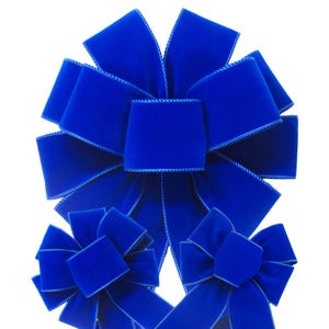 May include: A large, vibrant royal blue velvet bow with a central knot and multiple radiating loops, accompanied by two smaller matching royal blue velvet bows positioned below it, all featuring a distinct stitched edge detail.