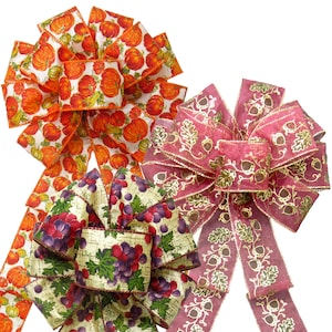 May include: Three different fabric ribbon bows in fall colors and patterns. The first bow is orange and white with a pumpkin pattern. The second bow is red with a gold acorn and leaf pattern. The third bow is green and white with a grape pattern.
