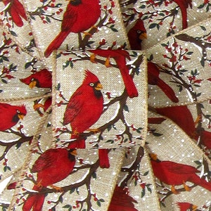 May include: Red cardinal birds perched on branches with green leaves and red berries against a beige burlap background with white flecks resembling snow.