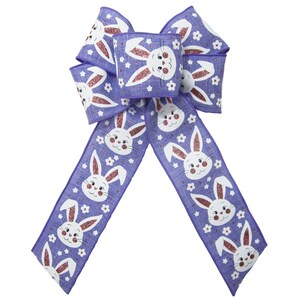 May include: A purple Easter bow with a repeating pattern of white bunny faces with pink glitter accents and small white flowers. The fabric bow has long tails. Ideal for Easter decorations.