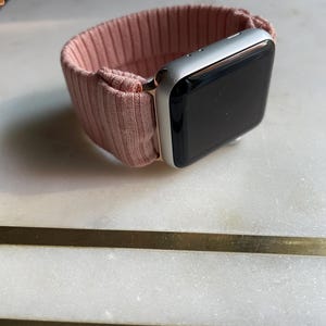May include: A silver smartwatch with a black screen and a blush pink ribbed fabric band. The band is secured to the watch with rose gold-colored hardware. The watch is resting on a white surface.