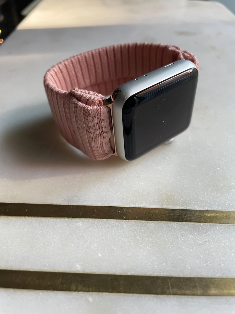 May include: A silver smartwatch with a black screen and a blush pink ribbed fabric band. The band is secured to the watch with rose gold-colored hardware. The watch is resting on a white surface.