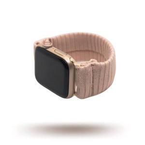 May include: A gold-colored smartwatch with a black screen and a blush pink ribbed knit band. The band appears to be made of a soft, stretchy material. The watch is shown floating against a white background.