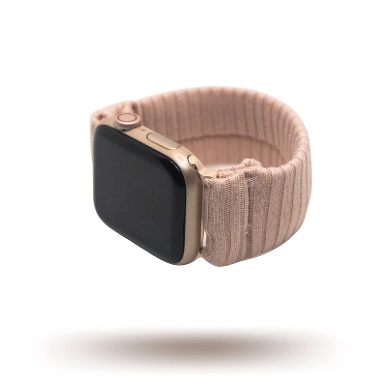 May include: A gold-colored smartwatch with a black screen and a blush pink ribbed knit band. The band appears to be made of a soft, stretchy material. The watch is shown floating against a white background.