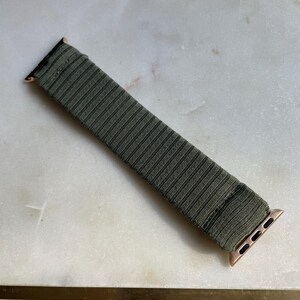 May include: Olive green Apple Watch strap with a ribbed texture. The strap has rose gold connectors. The strap is on a white and grey marble surface. The strap is a replacement for a smart watch.