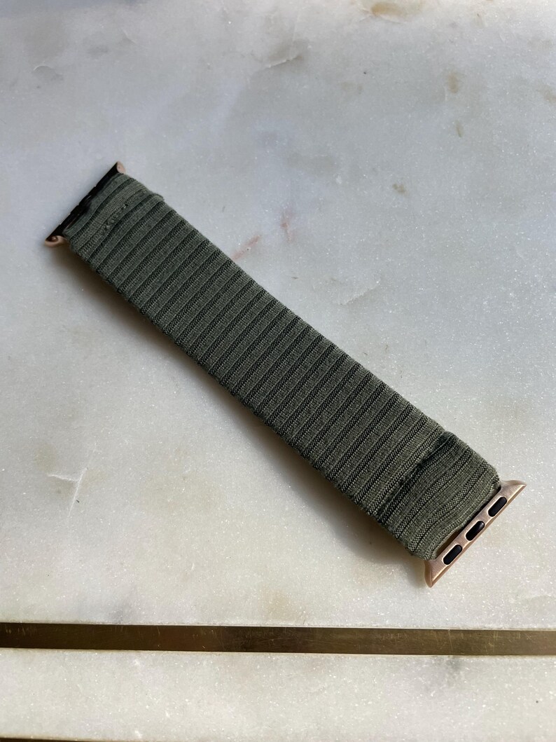 May include: Olive green Apple Watch strap with a ribbed texture. The strap has rose gold connectors. The strap is on a white and grey marble surface. The strap is a replacement for a smart watch.