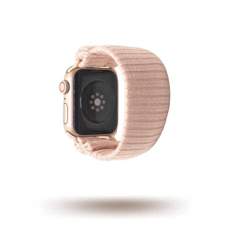 May include: A gold-colored smartwatch with a light pink ribbed fabric band. The watch face is black, and the band is looped around the watch. The watch is shown against a white background.