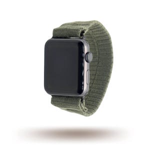 May include: A black smartwatch with a dark green ribbed fabric strap. The watch features a rectangular screen and a silver side button. The strap is made of a soft, flexible material, and the watch is suspended in the air.
