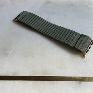 May include: An olive green and black striped watch strap with rose gold connectors. The strap is made of a textured fabric and is designed for a smartwatch. The strap is on a white marble surface.