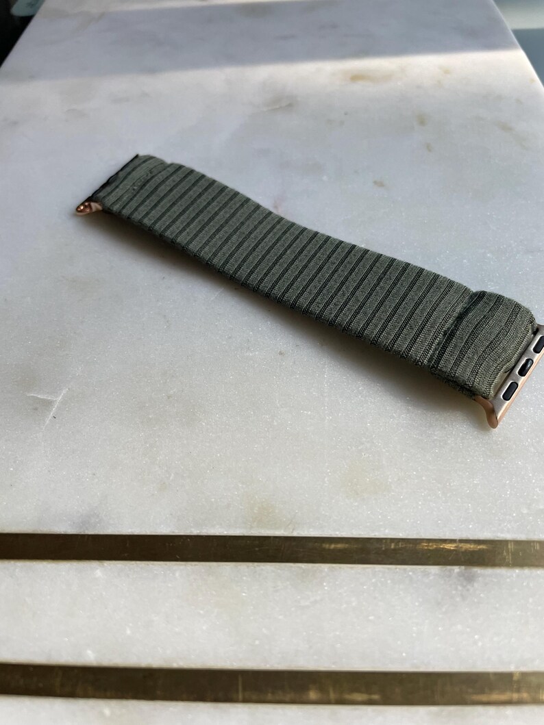 May include: An olive green and black striped watch strap with rose gold connectors. The strap is made of a textured fabric and is designed for a smartwatch. The strap is on a white marble surface.