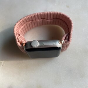 May include: A silver smartwatch with a pink ribbed fabric band. The watch face is black, and the band is looped around the watch. The watch has a digital display and a side button.