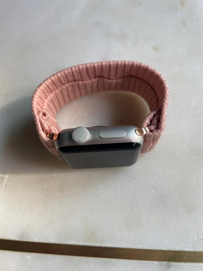 May include: A silver smartwatch with a pink ribbed fabric band. The watch face is black, and the band is looped around the watch. The watch has a digital display and a side button.