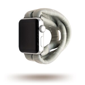 May include: Silver smartwatch with a black screen and a light gray fabric band. The band has a subtle grid pattern. The watch is suspended in the air against a white background, with a shadow below.