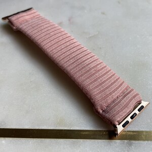 May include: A blush pink watch band with a ribbed texture. The band has gold-colored connectors on each end. The watch band is lying on a white and gray marble surface.