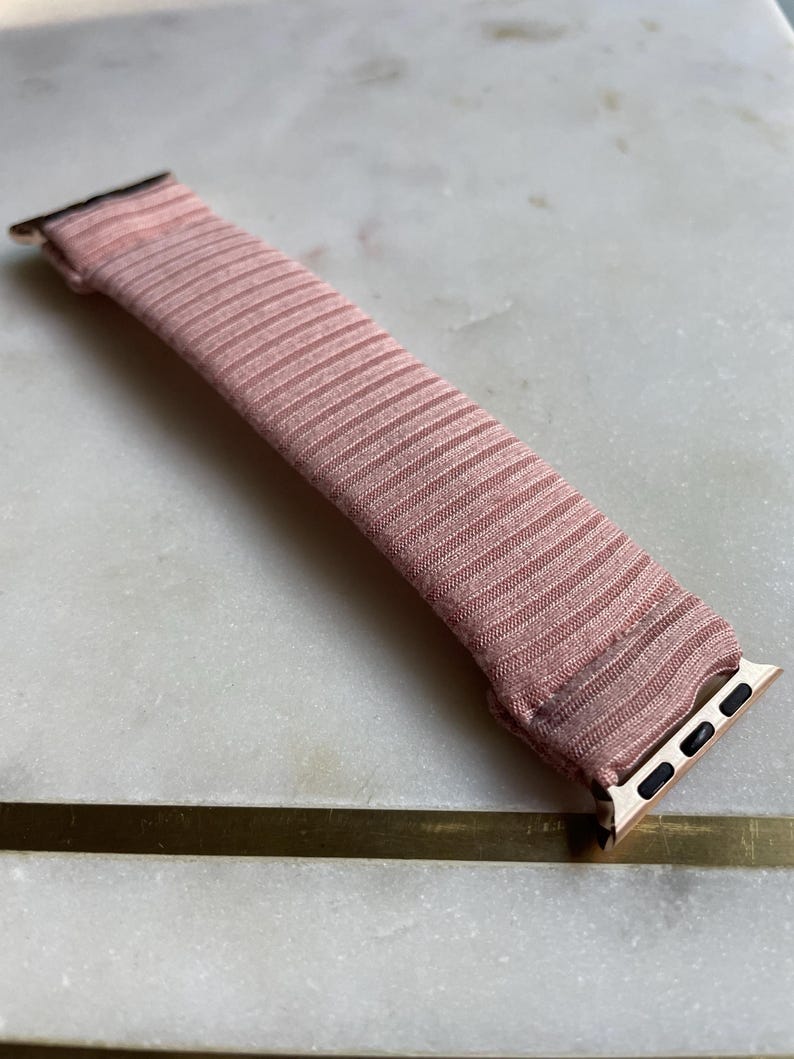 May include: A blush pink watch band with a ribbed texture. The band has gold-colored connectors on each end. The watch band is lying on a white and gray marble surface.