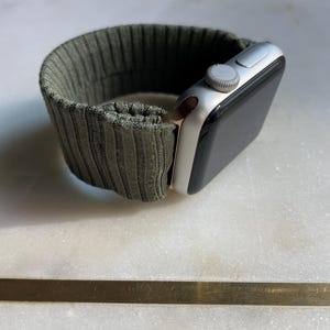 May include: Silver smartwatch with a dark olive green ribbed fabric strap. The watch face is black, and the strap is secured with a silver clasp. The watch is displayed on a white marble surface.