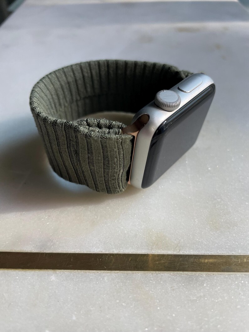 May include: Silver smartwatch with a dark olive green ribbed fabric strap. The watch face is black, and the strap is secured with a silver clasp. The watch is displayed on a white marble surface.
