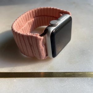 May include: A silver smartwatch with a black screen and a light pink ribbed fabric band. The band is looped around the watch face, showcasing the design and texture of the material. The watch is angled, displaying the side and top.