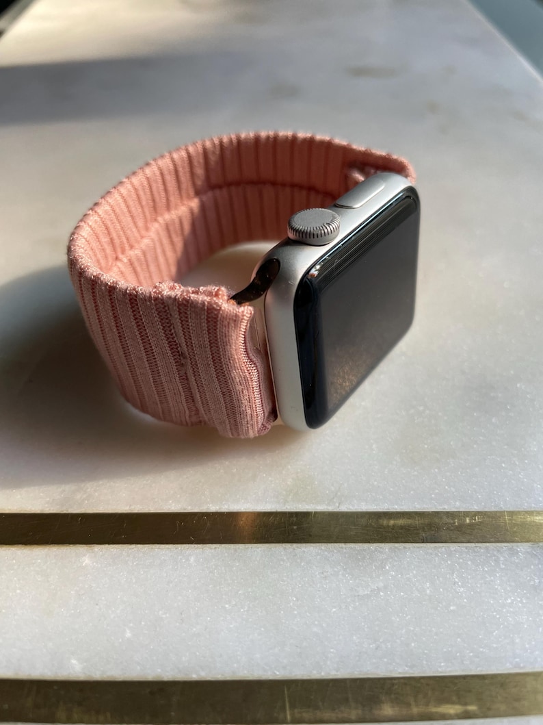 May include: A silver smartwatch with a black screen and a light pink ribbed fabric band. The band is looped around the watch face, showcasing the design and texture of the material. The watch is angled, displaying the side and top.