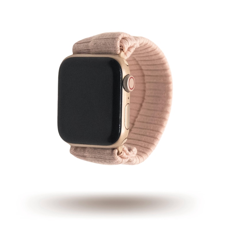 May include: A gold-colored smartwatch with a black screen and a blush pink fabric band. The band appears to be made of a soft, textured material. The watch is shown against a white background.