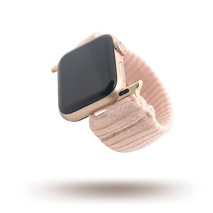 May include: A gold-colored smartwatch with a black screen and a blush pink fabric band. The watch is shown floating in the air, with a shadow beneath it. The band is a stretchy, ribbed material.