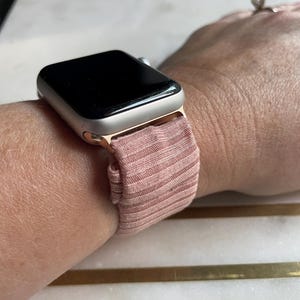 May include: A rose-colored Apple Watch with a matching ribbed fabric band is shown on a wrist. The watch face is black and the case is a light gold color. The band appears to be made of a soft, stretchy material.
