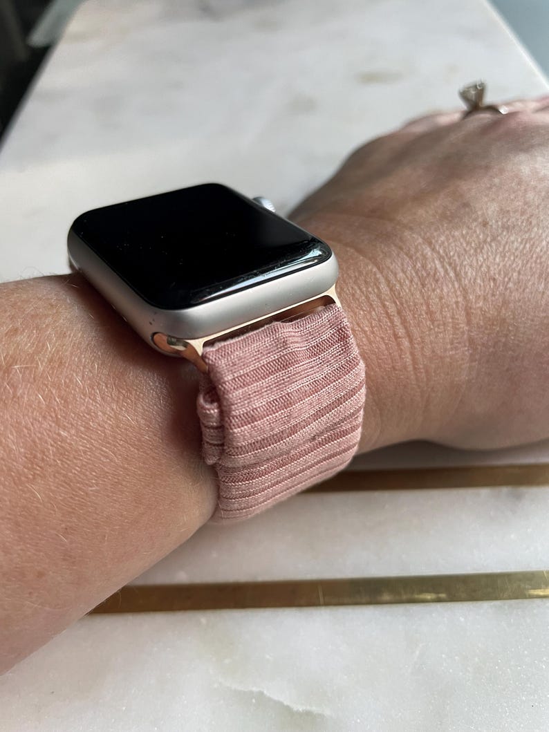 May include: A rose-colored Apple Watch with a matching ribbed fabric band is shown on a wrist. The watch face is black and the case is a light gold color. The band appears to be made of a soft, stretchy material.