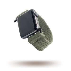 May include: A dark grey smartwatch with a green ribbed fabric strap. The watch face is black and the strap is a muted olive green. The watch is shown against a white background.