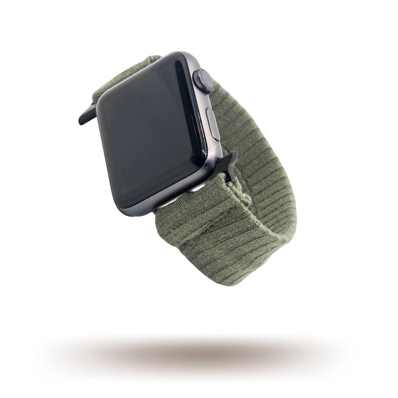 May include: A dark grey smartwatch with a green ribbed fabric strap. The watch face is black and the strap is a muted olive green. The watch is shown against a white background.