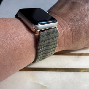 May include: A silver smartwatch with a black screen and olive green striped strap is shown on a wrist. The watch case has a gold finish. The strap is made of a textured fabric. The watch is a modern accessory.