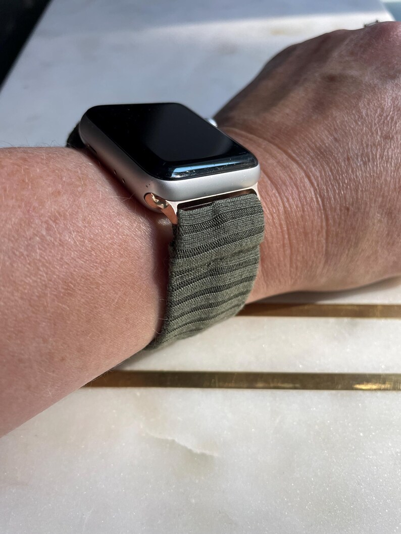 May include: A silver smartwatch with a black screen and olive green striped strap is shown on a wrist. The watch case has a gold finish. The strap is made of a textured fabric. The watch is a modern accessory.