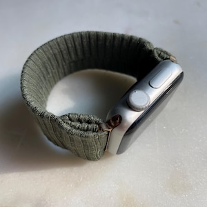 May include: Silver smartwatch with an olive green ribbed fabric strap. The watch face is black with a silver dial and button. The strap has a textured, ribbed design.