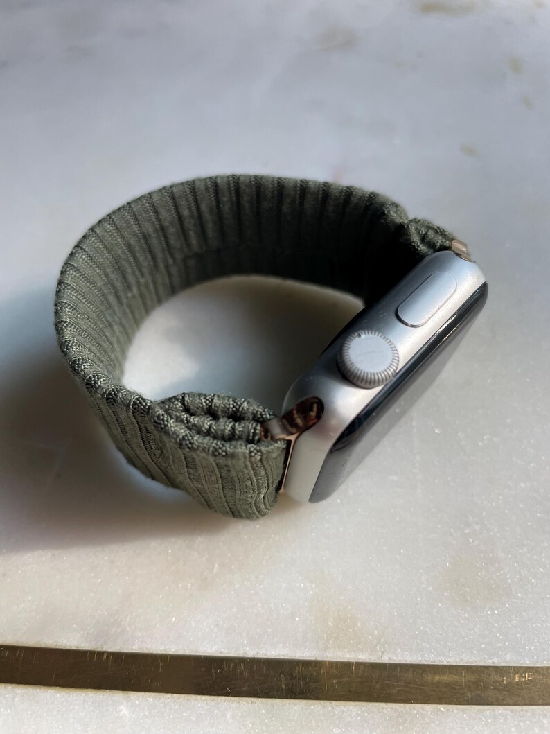 May include: Silver smartwatch with an olive green ribbed fabric strap. The watch face is black with a silver dial and button. The strap has a textured, ribbed design.
